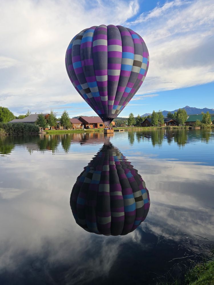 Hot air balloon with colorful stripes reflected in a calm lake with houses and trees in the background.