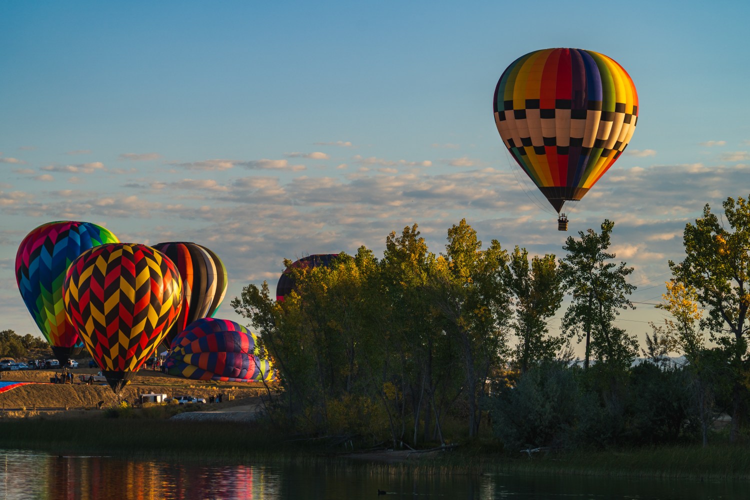 Colorful hot air balloons above trees by a lake during sunset.