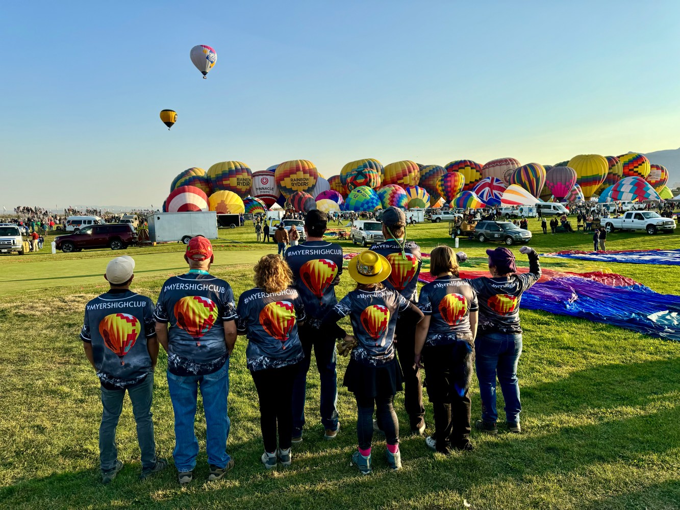 People wearing matching shirts watch hot air balloons on a grassy field under a clear sky.
