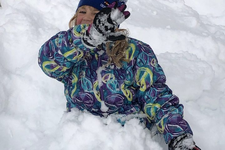 Child in colorful jacket plays in snow, wearing blue hat and gloves, partially buried in snow.